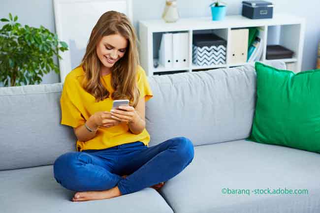 Portrait of beautiful young woman looking at phone and smiling while sitting on couch at home