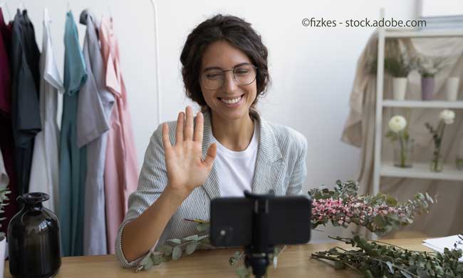 Smiling young Caucasian florist blogger wave at camera shooting floral composition arrangement on cell camera